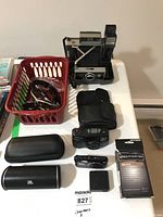 View of lot on white table: Polaroid 430 Land Camera with flash, Nikon Coolpix S3700 camera with case, Pentax Espoo 738G film camera, JBL cylindrical Bluetooth speaker, 3-in-1 Metro tuner in box, red basket with sunglasses, camera cases.