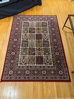 Full view of carpet showing the rectangular, multicolored patchwork design and red floral border laid on hardwood floor.