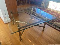 Coffee table with clear squared glass top and gold-toned metal base showing table from a side angle on hardwood floor.
