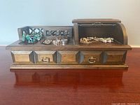 Front view of wooden jewelry box with assortment of costume jewelry on top and inside compartment.