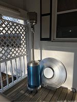 Full view of standing blue and stainless steel Crown Verity patio heater with large round reflector dish leaning against the wall on a wooden deck.