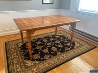 Full view of oak dining table from a corner angle showing turned legs and wood grain top on a patterned black and beige rug.