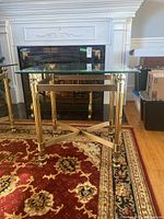 Front angle of the end table showing metal base and glass top, placed on a patterned rug in front of a fireplace.