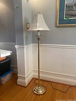 Full view of brass floor lamp with white shade standing on wooden floor near wall.