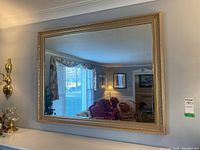 Framed mirror hanging on a wall with room reflection showing furniture and windows.