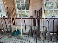 Full view of all gardening tools including shovels, rakes, rotary cultivator, hoes and forks aligned against a wall on wooden floor inside a room with windows.