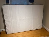 Front view of white IKEA double mattress standing upright against a wall on wood floor, showing vertical quilting.