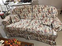 Front view of floral upholstered love seat showing curved backrest, rolled arms, cushioned seat and matching pillows on the carpeted floor near white furniture.