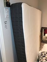 Full view of the queen size mattress standing upright showing white sleep surface and gray side panel with stitching detail.