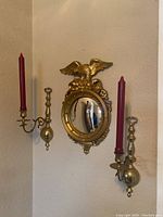 Front view showing both brass candle sconces mounted on wall with red candles, surrounding an oval fisheye mirror topped with a gold eagle frame.