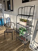 Full view of the black metal chair and metal plant stand with shelves, including faux plants and candles placed on shelves.
