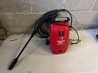 Front and side views of red and black HSDS power washer showing on/off switch, branding, power cord wrapped at back, and attached spray gun with extended lance nozzle.