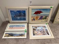 Five framed coastal and beach themed prints displayed on carpeted floor against white wall. Visible details of artwork and frames.