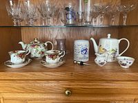 Shelf with fine china teapots, teacups, saucers, Blue Jays mug, and glassware on upper shelf