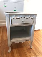 Front and side view of white wooden side table with one drawer and open shelf below, placed on hardwood floor.