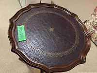 Top view of a small circular wood side table with a decorative leather top and scalloped wood edge, showing a gold floral pattern design.