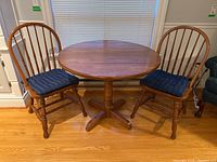 Full view of round drop leaf wood table with two wooden spindle back chairs, each with blue seat cushions.