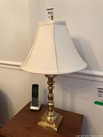 Full view of one brass lamp showing turned base and cream fabric shade alongside cordless phone for scale.