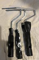 Three paint roller frames with black handles, two have white paint residue, metal roller frame visible, no roller covers included.