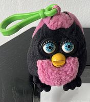 Close-up photo of a small black and pink plush Furby keychain with blue plastic eyes and yellow beak, attached to a green clip.