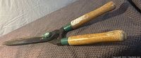 Photo showing a pair of garden shears with wooden handles and metal blades, placed on a textured fabric surface.