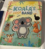 Front cover of the children's book 'Koala's Cool Band' showing colorful animals and a rubber koala figure holding a tambourine on the cover.