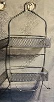 Metal shower caddy hanging on a textured wall, showing two mesh shelves and hooks for hanging