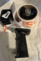 Black handheld tape dispenser with clear packing tape roll and extra black tape core on light colored fabric surface.