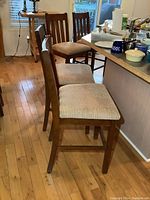 Four wooden bar height chairs shown in a row from the side, highlighting the seat upholstery and wooden frame, placed in a kitchen/dining area with hardwood floors.