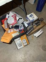 Photo showing the mixed lot of items including bright orange cleaning cloths, a boxed pest repeller, a black Ott Lite lamp, various boxed items including a pedicure kit, and a smoke detector in packaging.