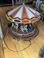 Carousel music box with visible horses, marquee canopy top, gold ornamentation, power cord on wooden floor