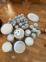 Large set of white German China pieces with gold trim including cups, plates, shallow bowls, saucers, and oval platters spread out on wooden floor.