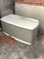 Beige Rubbermaid outdoor deck box viewed from the front left side, showing the body and lid with some surface dirt and minor wear.