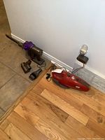 Dyson cordless vacuum with three attachments and Dirt Devil handheld vacuum on floor next to wall outlet.