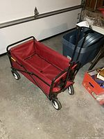 Side view of the folding wagon showing red fabric sides, black metal frame, and white plastic wheels on concrete floor.