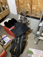 View of golf clubs in black golf bag standing on concrete floor with some boxes and ladder in background.