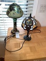 Both lamps side by side on wooden table with window blinds in background.