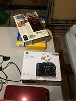 Boxed GE X500 digital camera and two boxed HD handheld digital camcorders on table.