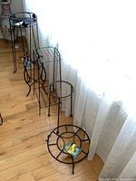 Photo showing all five metal plant stands of varying heights and designs, arranged on wooden floor near a window with lace curtains.