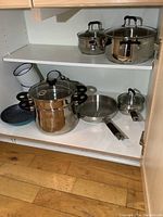 Stainless steel and enamelware cookware displayed inside a cabinet on two shelves, showing various pots, pans, and enamel mugs and bowls.