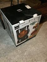 Boxed Vertigenius vertical rotisserie photographed on floor, showing images of rotisserie with cooked chicken and meals.