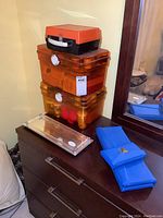 Stack of two large translucent orange plastic cases and one small black and orange case on a dark brown dresser, accompanied by two folded blue cases and a small clear plastic mirror.