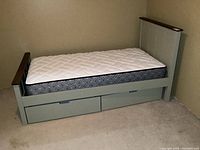 Full view of single bed with mattress and two large drawers underneath, wood frame in muted greenish-gray with dark wood trim on headboard and footboard.