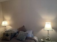 Pair of table lamps lit on either side of a bed with pillows against a beige wall.