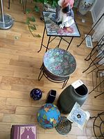 Metal framed table with colorful tile top featuring butterflies and flowers, porcelain floral bowl on stand, small vases, and taller floral vase on wooden floor.