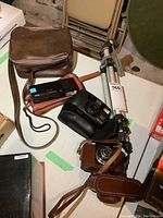 Lot overview photo showing multiple cameras in leather cases, a black Polaroid-style camera with strap, a brown leather carrying case, and a metallic tripod with adjustment knobs.