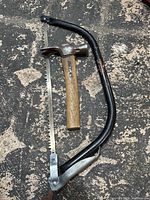 Photo showing the bow saw and hammer laid on a dark surface, highlighting the saw blade and hammer handle.