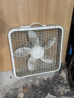 White box fan with metal front grill and four blades, sitting on the floor against wood paneling.
