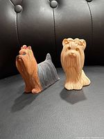 Pair of Don Jones Yorkshire Terrier dog figurines, one painted brown and gray with a red bow, the other cream-colored, photographed on black leather background.