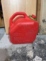 Red plastic gas can with embossed safety warnings, integrated handle, and green locking cap.